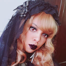 Aria with orange wavy hair and bangs. She has one dark makeup and is wearing a black rectangular headdress and veil.